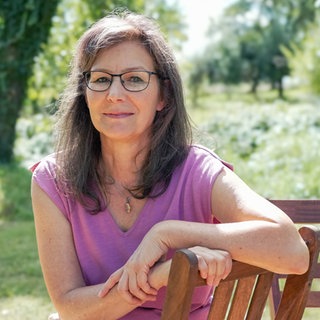 Tanja trägt eine Brille und lächelt schwach in die Kamera. Sie sitzt an einem Sommertag im Grünen auf einem Stuhl und spricht über den Tod ihres Sohnes und ihre Trauer.