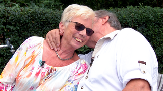 Das Paar Uschi und Sigi sitzt in Uschis Garten. Sigi hält Uschi im Arm und küsst sie liebevoll auf die Wange, während Uschi glücklich lacht. 