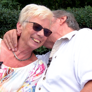 Das Paar Uschi und Sigi sitzt in Uschis Garten. Sigi hält Uschi im Arm und küsst sie liebevoll auf die Wange, während Uschi glücklich lacht. 