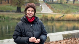 Alex steht lächelnd mit dicker schwarzen Daunenjacke und brauner Mütze vor dem See im Rosensteinpark. Ihre Augen sind gold geschminkt und sie trägt Ringe. 