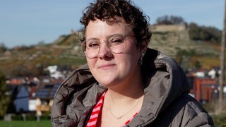 aro erhält mit Anfang 20 ihre Krebsdiagnose. Sie steht draußen, im Hintergrund sind Häuser zu sehen. Sie hat kurze gelockte Haare und schaut in die Kamera. 