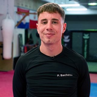 Patrick steht im Kickbox-Studio und lächelt in die Kamera. Er trägt ein schwarzes Trainingsshirt.