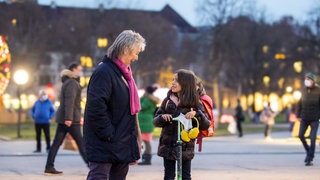 Adelheid und Emilia haben warme Winterkleidung an und stehen auf einem Platz. Emilia hat ihren Cityroller dabei und die zwei unterhalten sich lächelnd.