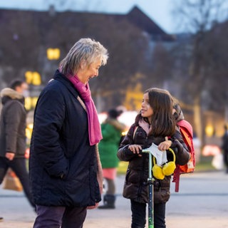 Adelheid und Emilia haben warme Winterkleidung an und stehen auf einem Platz. Emilia hat ihren Cityroller dabei und die zwei unterhalten sich lächelnd.