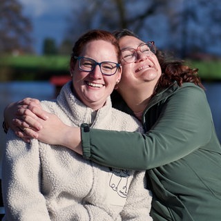 Anky (links) und Conny (rechts) sind beste Freundinnen und leben beide mit MS. Sie sind draußen und Conny umarmt Anky. Anky lacht in die Kamera. 