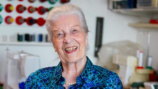 Dietlinde sitzt in ihrer Schneider-Werkstatt in ihrer Wohnung und lächelt in die Kamera. Hinter ihr an der Wand hängt Nähgarn, in Kisten stapeln sich Stoffe und Knöpfe und hinter ihr steht eine ihrer Nähmaschinen. 