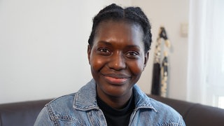 Grace hat schwarze Haut, ihre schwarzen Haare sind dich am Kopf geflochten. Sie trägt einen Jeans-Overall und lächelt in die Kamera. 