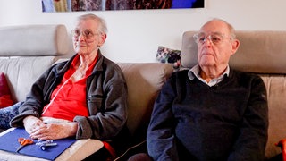 Roland Sing und seine Frau Ursula sitzen auf ihrer beigen Couch in ihrem Wohnzimmer, hinter ihnen an der Wand hängt ein Bild. Ursula, die Long-Covid und eine Lungenfibrose hat, trägt eine Sauerstoffbrille und auf ihrem Schoß liegen ihre Bastel-Utensilien. 