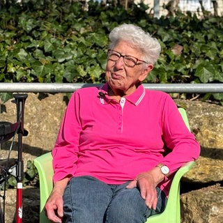 Hildegard sitzt im Garten der Tagespflege in Bad Cannstatt. Die Sonne scheint, sie trägt ein pinkes Polo und eine Brille. Ihr Rollator steht neben ihr, sie sitzt auf einem Gartenstuhl und lächelt.