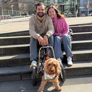 Leonie und Marvin sitzen auf einer Steintreppe und lächeln in die Kamera. Vor den beiden steht Hündin Lilly auf ihren Vorderbeinen. Ihr Rollstuhl hält ihre Hinterbeine.