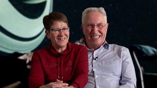 Sigrid und Joachim sitzen nebeneinander und lachen in die Kamera. Im Hintergrund ist verschwommen der Innenraum des Stuttgarter Planetariums zu sehen.