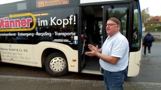 Isil trägt ein kurzärmeliges, hellblaues Hemd, und trägt eine braune Kappe. Er steht vor seinem Bus und wartet auf Fahrgäste. 