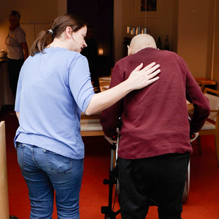 Melanie (links) arbeitet als Pflegerin im Hospiz. Sie läuft mit einem Gast (rechts) zum Esstisch, die beiden sind von hinten zu sehen. Melanies Arm liegt auf dem Rücken des Mannes.