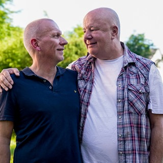 Ehepaar Ralf und Michael sind seit 37 Jahren ein Paar. Sie stehen Arm in Arm vor einer grünen Hecke und schauen sich tief in die Augen.
