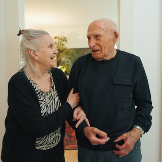 Josef pflegt mit 104 Jahren seine Frau Mira nach einem Sturz. In ihrer Wohnung stehen die beiden beieinander, Mira hat sich bei Josef am Arm eingehakt. Beide blicken lächelnd in die Kamera.