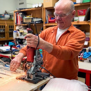 Klaus-Peter steht in einem orangenen Pullover und mit einer Schutzbrille in seiner Werkstatt und fräst ein Metallstück zurecht.  