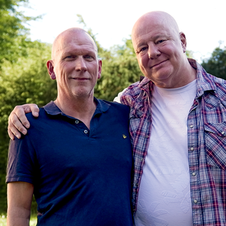 Michael (rechts) und Ehemann Ralf (links) stehen vor Bäumen in einer Wohnsiedlung. Das Ehepaar, beide mit Glatze, halten sich im Arm und lächeln in die Kamera. 