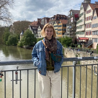 Laura steht auf der Eberhardsbrücke in Tübingen. Sie hat eine Jeansjacke an und ihre Hände in den Taschen. Hinter ihr sieht man Tübingen mit dem Hölderlinturm. 