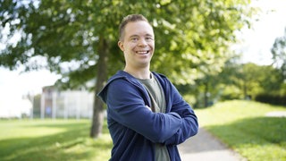 Daniel lebt in einer inklusiven WG. Er steht im freien, verschränkt die Arme und lächelt in die Kamera.