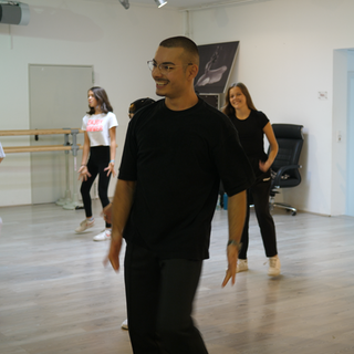 Tanzlehrer Deniz ist in einem Tanzstudio. Dort bringt er Kindern das Tanzen bei. Deniz macht eine Tanzübung vor und lacht dabei. Im Hintergrund sind Kinder zu sehen, die Deniz’ Tanzbewegung nachmachen. 