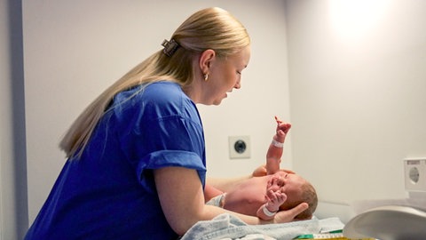 Pauline ist 27 Jahre alt und arbeitet als Hebamme in der Nachtschicht. Sie steht auf der linken Seite des Bildes und hält ein kleines Baby in der Hand, das auf einem Wickeltisch liegt.