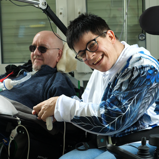 Im Vordergrund sitzt Nicole in ihrem Rollstuhl, sie hat kurzes schwarzes Haar und lächelt. Im Hintergrund liegt Sandro in einem Pflegebett und lächelt ebenfalls. Er trägt eine Sonnenbrille, Glatze und einen kurzen Bart.