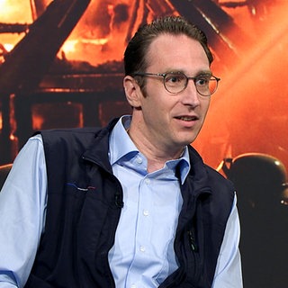 Andreas erzählt von seinem ehrenamtlichen Engagement und seiner Auszeichnung mit dem Verdienstorden. Im Hintergrund ist ein Foto von einem großen Brand mit Feuerwehrleuten zu sehen. 