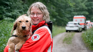 Nicole mit ihrer Hündin Cäthe auf dem Arm. Sie trägt eine orangene Einsatzjacke der Rettungshundestaffel.