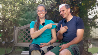  Das Paar Raphaela und Cornelius lacht herzlich miteinander auf einer Bank draußen im Freien. Die beiden haben zusammen sechs Söhne, eine richtige Großfamilie.