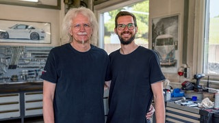 Marcel und sein Papa Rainer stehen im Familienbetrieb. Im Hintergrund sieht man Werkzeug. Bilder von Autos hängen an den Wänden. Marcel hat die Autowerkstatt übernommen. 