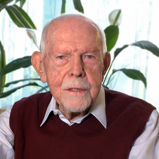 Helmut ist 95 Jahre alt, sitzt auf seinem Sofa und blickt in die Kamera.