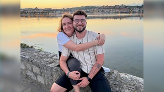 Julia und Jonathan sind seit 2015 ein Paar. Die beiden sind im Urlaub und sitzen auf einer Mauer. Im Hintergrund geht die Sonne unter und ein Gewässer ist zu sehen. Julia umarmt Jonathan von der Seite, beide lachen in die Kamera.