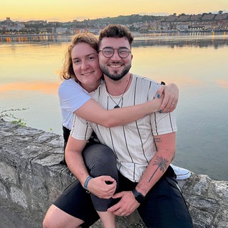 Julia und Jonathan sind seit 2015 ein Paar. Die beiden sind im Urlaub und sitzen auf einer Mauer. Im Hintergrund geht die Sonne unter und ein Gewässer ist zu sehen. Julia umarmt Jonathan von der Seite, beide lachen in die Kamera.