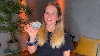 Maike hat Synästhesie. Sie sitzt in einem Studio auf einem Hocker. In der Hand hält sie ein Modell ihres Gehirns. Sie lächelt in die Kamera-