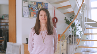 Maria hat ihre Tochter verloren – sie setzt sich dafür ein, dass Menschen auch auf der Arbeit Unterstützung beim Trauern bekommen. Maria steht bei sich zu Hause im Flur. Sie hat die Hände hinter dem Rücken und schaut in die Kamera. 