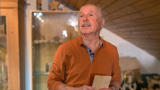 Armin steht vor einer Wand aus Holz, wo auch einige seiner Schnitzereien zu sehen sind. Er hat ein Holzbrett in der Hand und schaut neben der Kamera vorbei. 