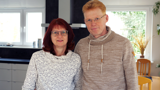 Ulrike und Gerold sind weiße Menschen in ihren 50ern. Sie trägt eine Brille und hat rötlich gefärbte Haare. Er trägt eine randlose Brille und hat hellrotes Haar.  