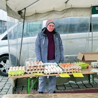 Susanne steht an einem Marktstand mit Eiern in Eierkartons. Sie trägt eine Wollmütze und warme Jacke und schaut leicht kritisch in die Kamera.