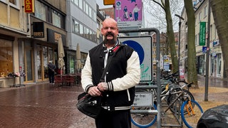 Steve ist ein großer Mann mit ausdrucksvollem Schnauzbart, Glatze und vielen Tätowierungen.
