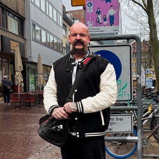 Steve ist ein großer Mann mit ausdrucksvollem Schnauzbart, Glatze und vielen Tätowierungen.