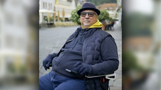 Ralf trägt einen Hut, eine Sonnenbrille und Handschuhe und sitzt in seinem Rollstuhl auf dem Schwäbisch Gmünder Marktplatz. Er lächelt in die Kamera.  