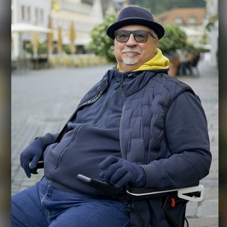 Ralf trägt einen Hut, eine Sonnenbrille und Handschuhe und sitzt in seinem Rollstuhl auf dem Schwäbisch Gmünder Marktplatz. Er lächelt in die Kamera.  