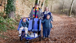 Eine Familie spaziert mit einem blauen Rehabuggy im Wald. Die Familie besteht aus Opa Peter, er trägt einen bunten Schal und Hut, Ex-Schwiegertochter Corinna, sie trägt einen weißen Schal und eine blaue Jacke und den Kindern Niklas und Damian, die eine Behinderung haben. Damian sitzt im Buggy und ist angeschnallt, Niklas wird von seiner Mama festgehalten, da beide kein Gefahrenbewusstsein haben und sie deshalb nicht allein weglaufen dürfen. 