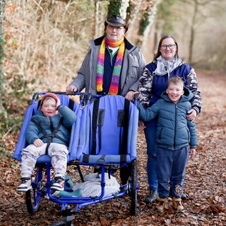 Eine Familie spaziert mit einem blauen Rehabuggy im Wald. Die Familie besteht aus Opa Peter, er trägt einen bunten Schal und Hut, Ex-Schwiegertochter Corinna, sie trägt einen weißen Schal und eine blaue Jacke und den Kindern Niklas und Damian, die eine Behinderung haben. Damian sitzt im Buggy und ist angeschnallt, Niklas wird von seiner Mama festgehalten, da beide kein Gefahrenbewusstsein haben und sie deshalb nicht allein weglaufen dürfen. 
