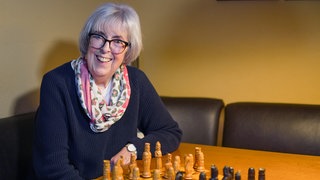 Als ihr Mann 1975 vier Wochen in der Kur ist, lernt Hanne das Schachspielen. Seitdem hat sie der Sport nicht mehr losgelassen. Hanne sitzt an einem Tisch vor dem Schachbrett. Sie lacht in die Kamera.