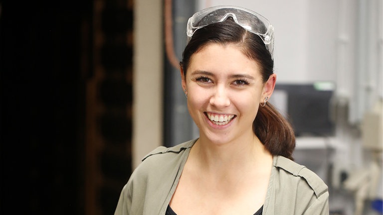 Junge Frau steht mit Schutzbrille und Bohrmaschine in einer Werkstatt und lächelt in die Kamera