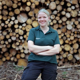 Eine junge Frau steht vor aufgestapeltem Holz.