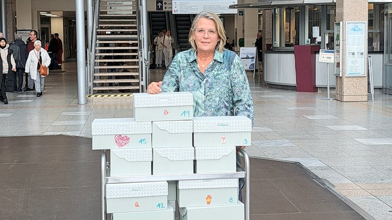 Charlotte steht im Eingangsbereich einer Klinik. Vor ihr steht ein Rollwagen, auf dem einige Kisten gestapelt sind.