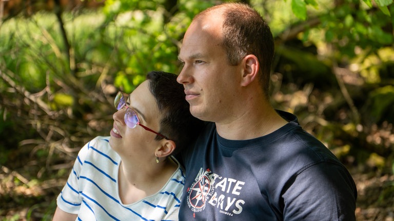 Ein Mann im dunkelblauen T-Shirt und eine Frau im gestreiften T-Shirt sitzen auf einer Bank im Grünen.
