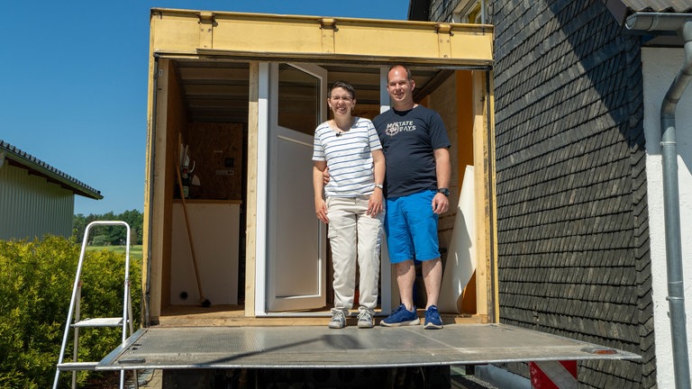 Eine Frau im gestreiften T-Shirt mit heller Hose und ein Mann im dunkelblauen T-Shirt mit kurzer Hose stehen auf der Ladefläche eines LKWs.